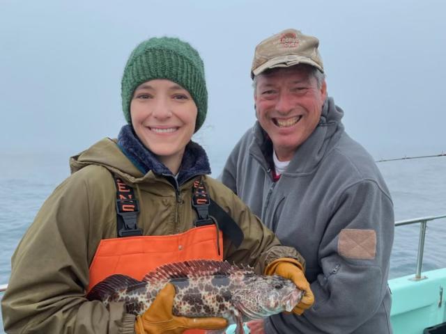 Two anglers standing on a boat, one of them holding a large brown and black lingcod.