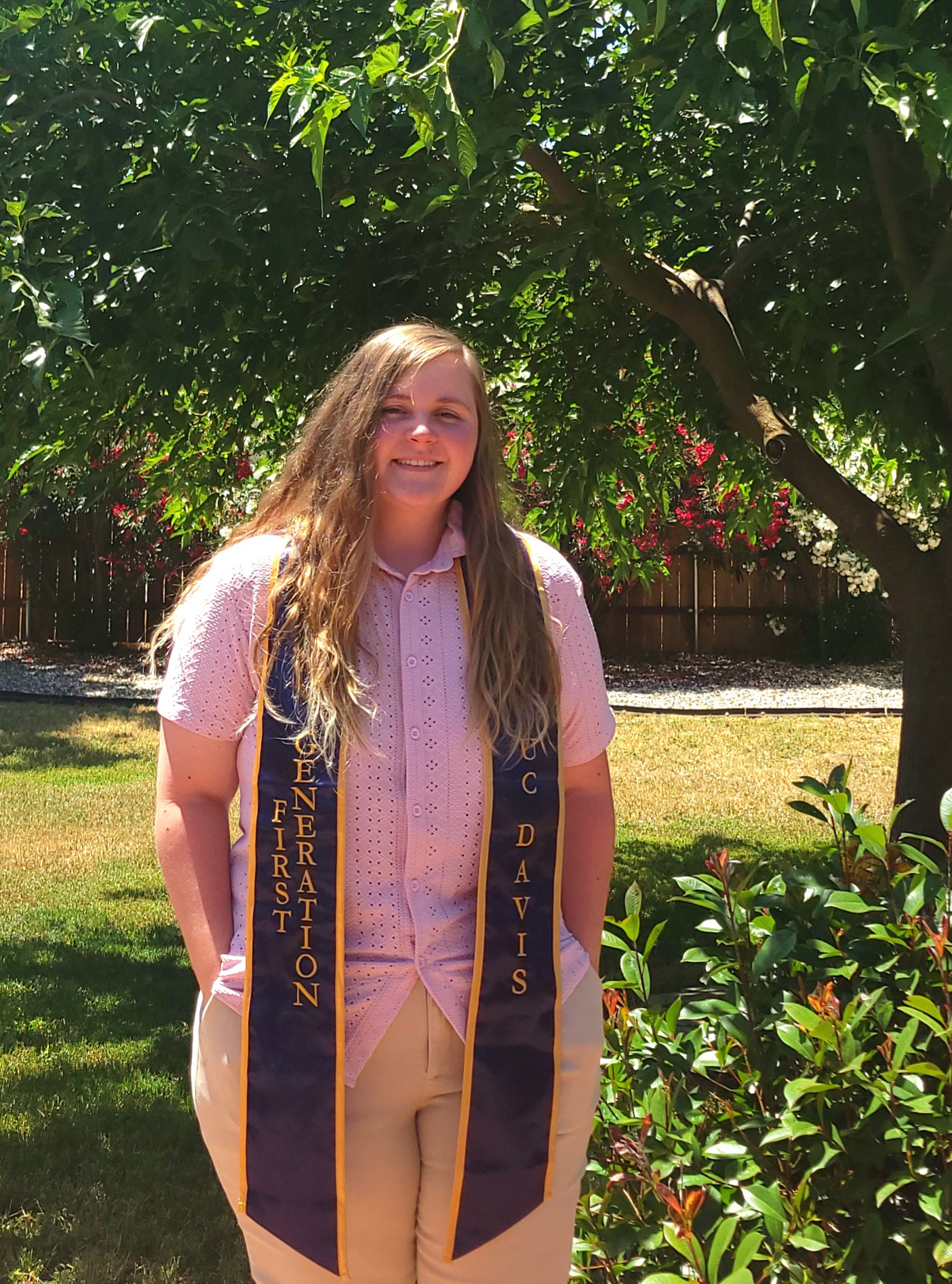 A graduation photo of Oceanna Warnock, wearing a white shirt and gold and blue UC Davis stole, posing in front of background greenery.