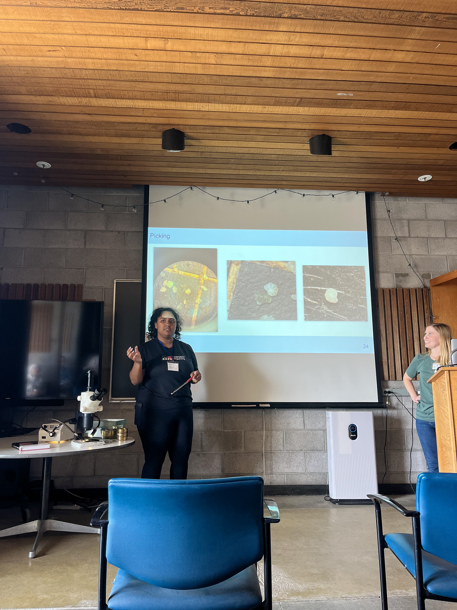 Two presenters in wooden-ceiling room with projected slides and blue chairs