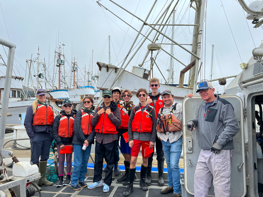 A group of students in bright life jackets standing on a dock surrounded by boats.