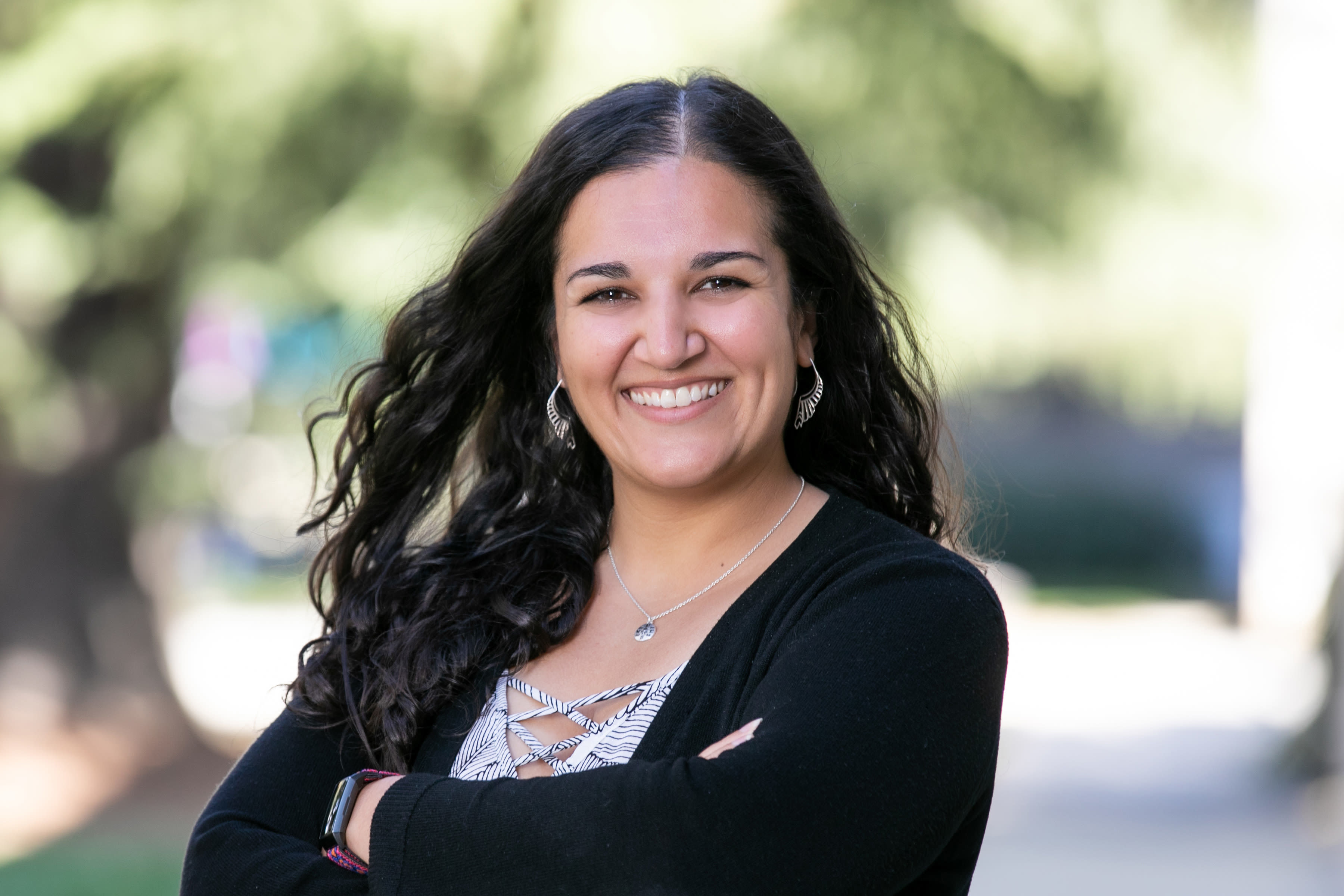 Outdoor headshot of smiling woman with long dark hair, arms crossed, blurred trees