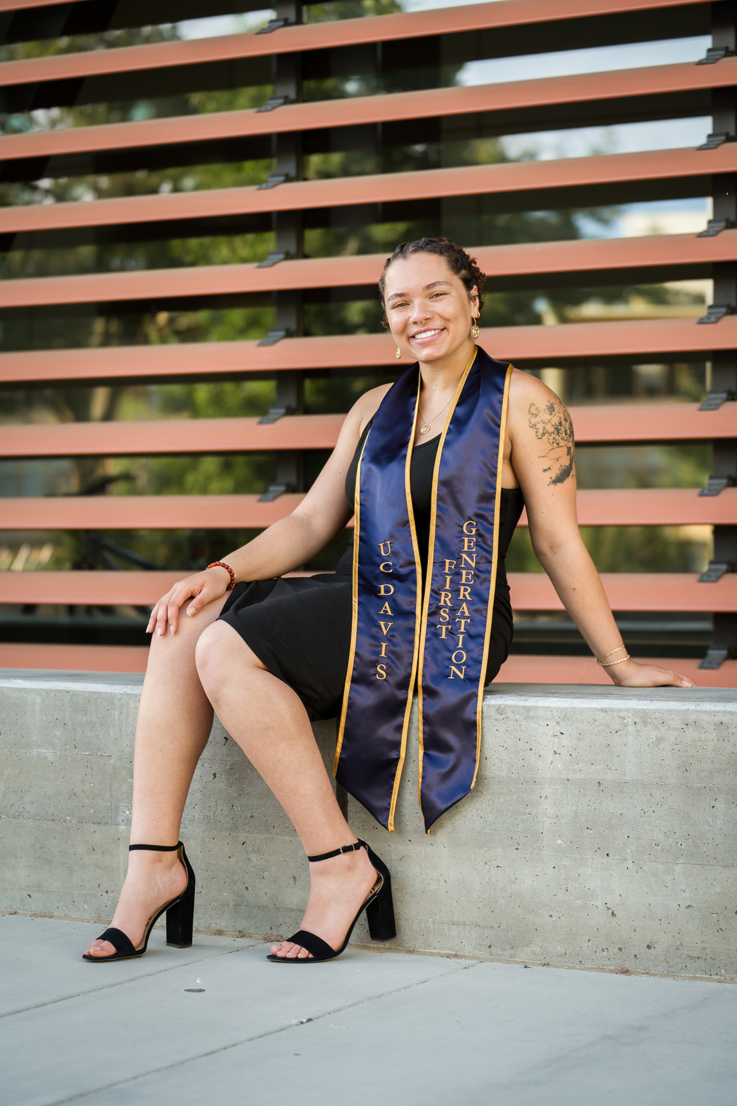 A photo of Genece Grisby, wearing a black dress and a blue and gold UC Davis stole, sitting on a cement step and smiling at the camera