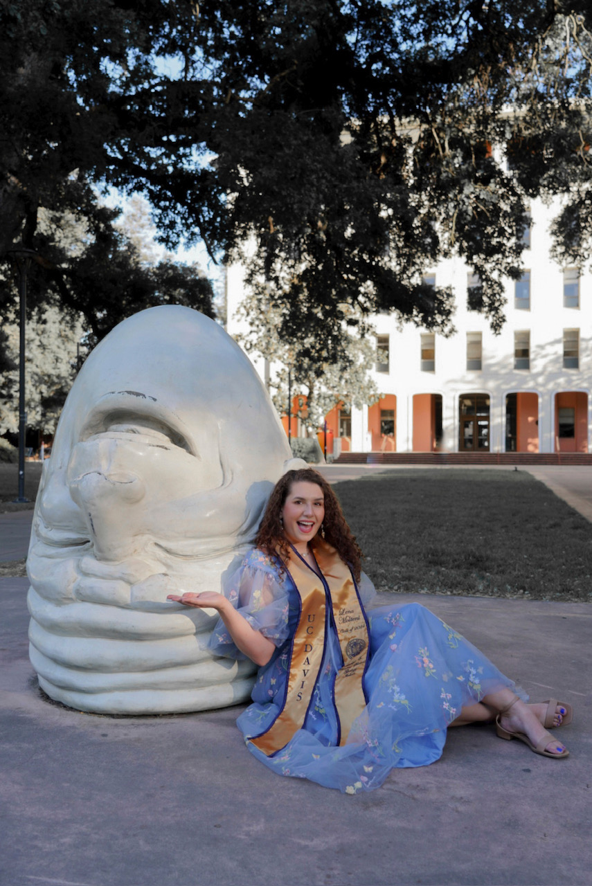 A graduation photo of Lena Molteni, who is wearing a blue gown, gold and blue UC Davis stole, and posing next to one of the UC Davis egghead statues.
