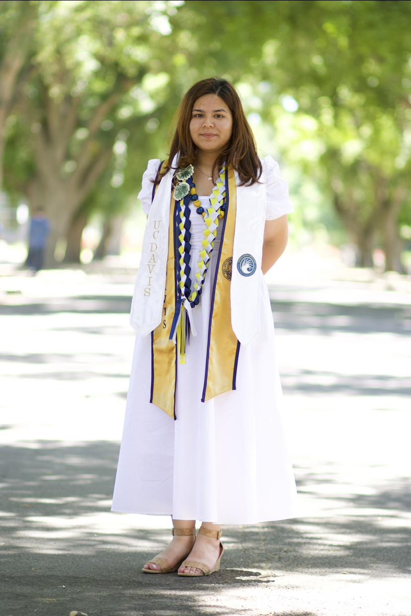 A graduation photo of Jade Valdez, who is wearing a long white dress, brown sandals, and blue and gold UC Davis graduation stoles.