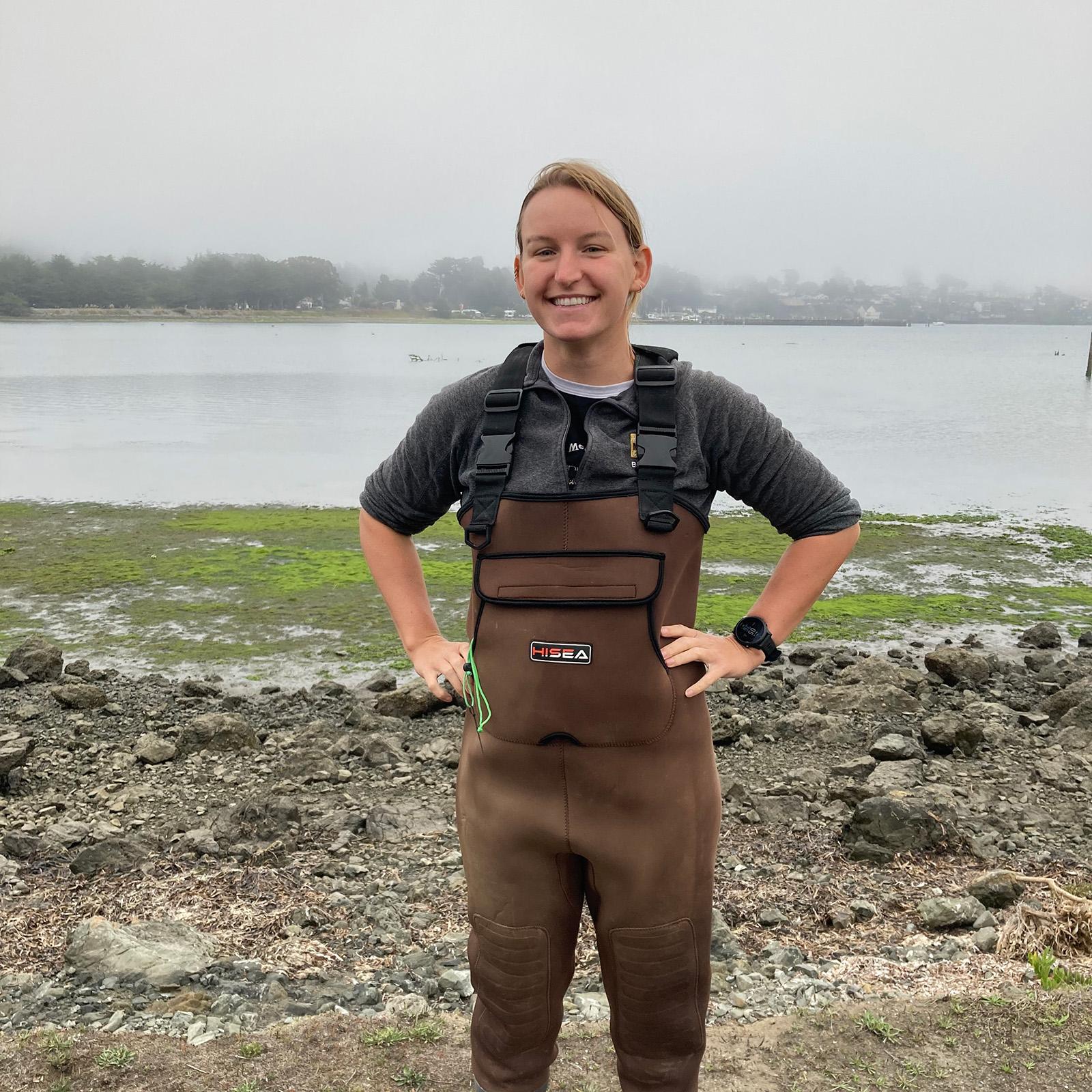 A portrait of Katie Erickson standing in a mudflat wearing waders
