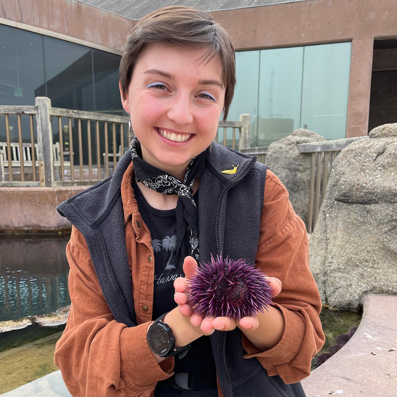 A portrait of Maddie Armstrong standing in front of BML and holding a purple sea urchin
