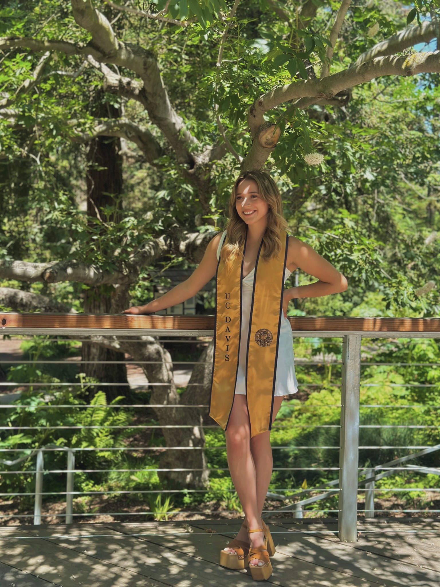 A photo of Sam Martinez, wearing a white dress and gold and blue UC Davis graduation shawl, standing in the shade under a leafy tree.