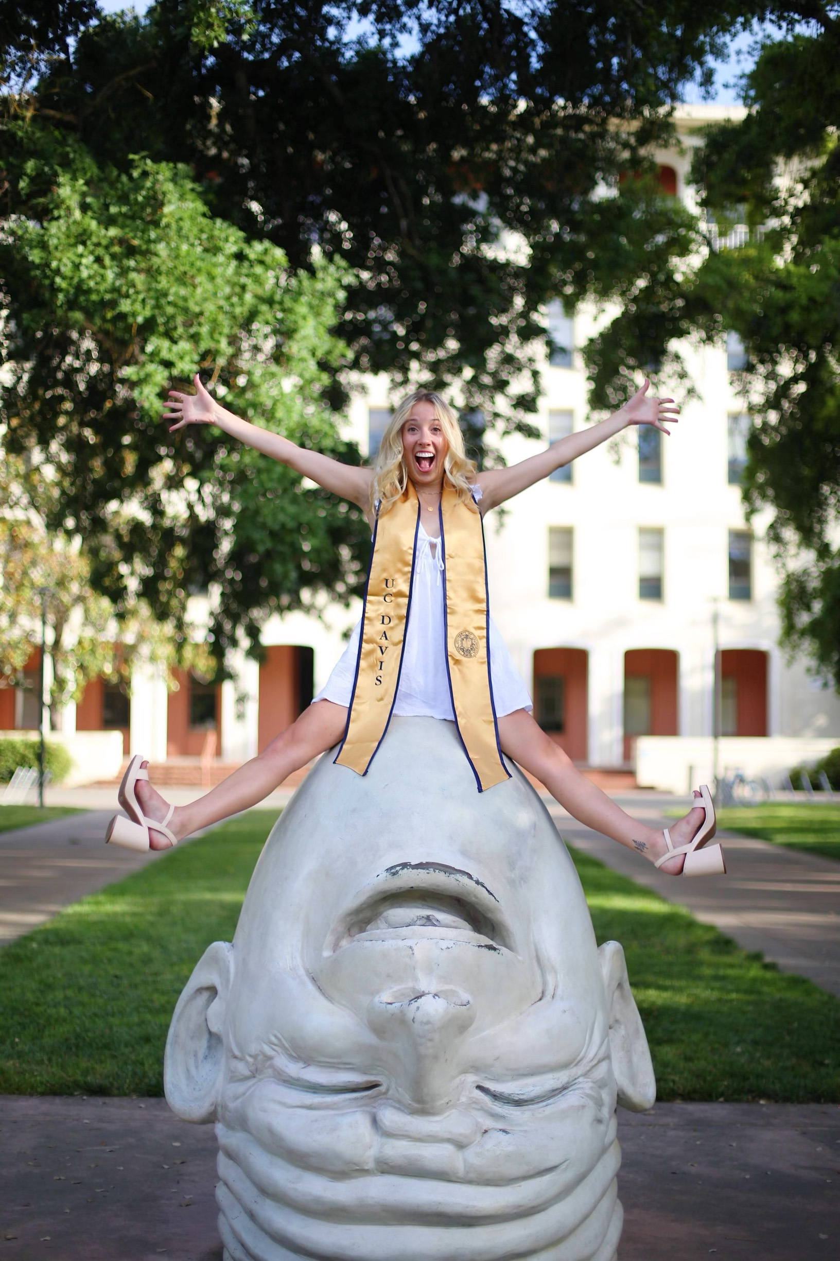 An image of Savannah Torgerson sitting atop one of the UC Davis Eggheads, wearing a white dress and gold and blue UC Davis graduation shawl, with a joyful and excited expression on her face.