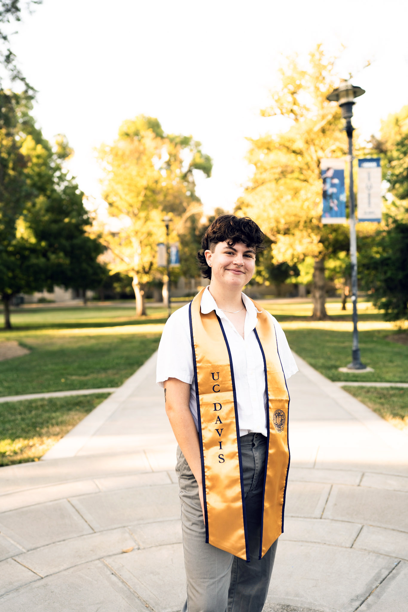 A graduation photo of Adri Penix, who is wearing a white shirt, khaki slacks, and a navy and gold UC Davis graduation stole, standing in front of a tree lined walkway.
