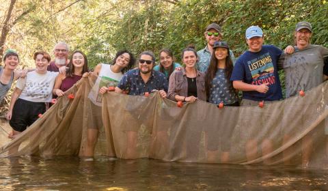 Group of people standing in water, holding a fishing net. Trees in the background.