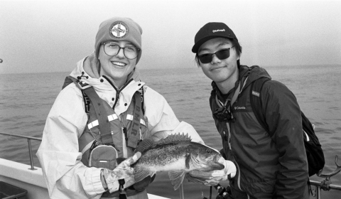 A black and white photo of SRJC Intern Alex Margand posing with another fisher, holding a large rockfish between them