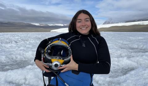A portrait of Abbey Dias, holding diving gear and standing in front of an expanse of snow.