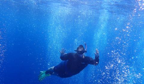 A full-body shot of Anya Brown snorkeling. She raises her hands up as bubbles swirl around her. 