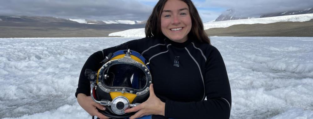 A portrait of Abbey Dias, holding diving gear and standing in front of an expanse of snow.