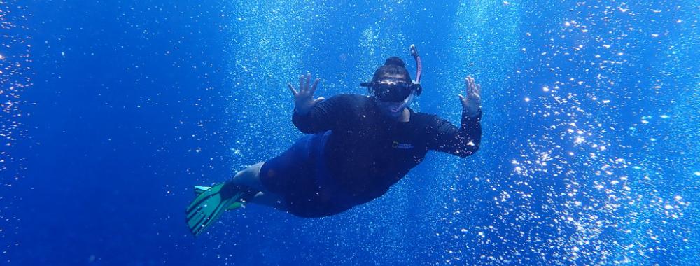 A full-body shot of Anya Brown snorkeling. She raises her hands up as bubbles swirl around her. 