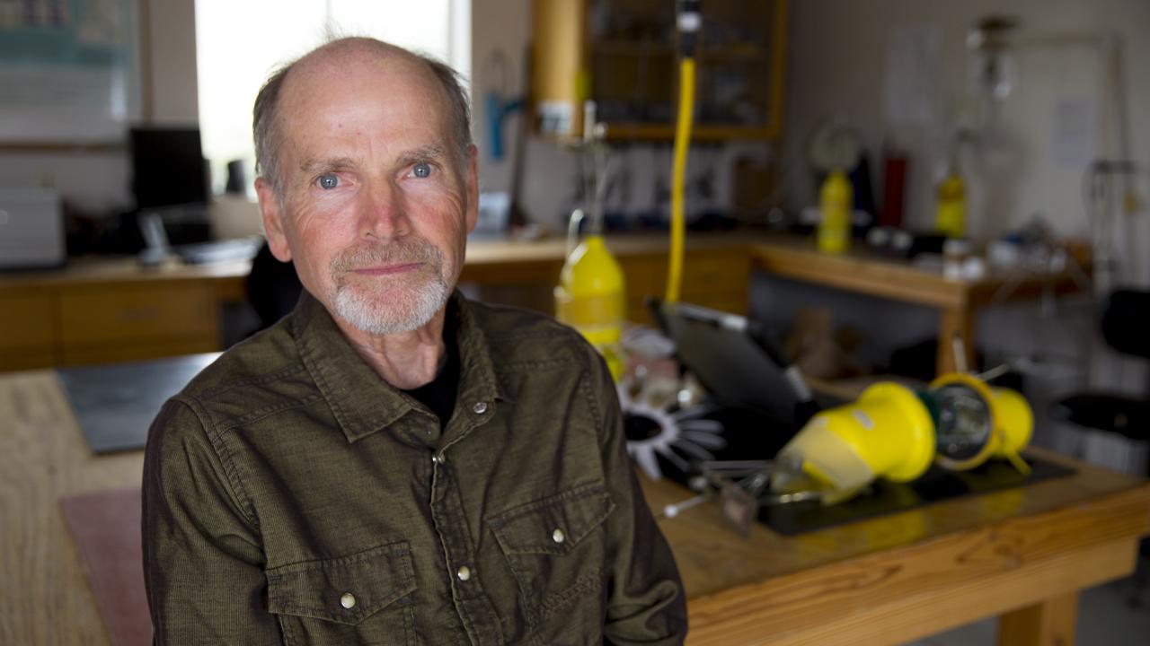 A person with gray hair and blue eyes, wearing a dark green hooded jacket, standing in front of a table that has bright yellow robot equipment on it.