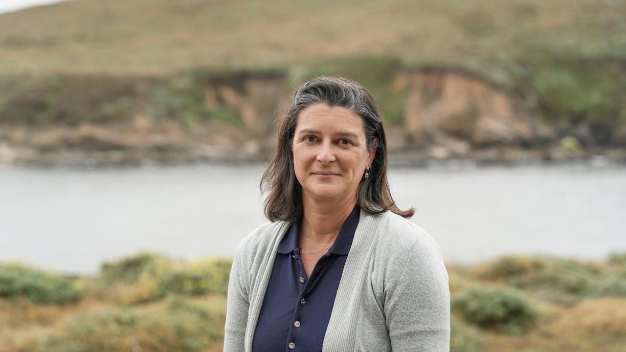 A portrait of Tessa Hill wearing a sweater and shirt and standing in front of a coastal background.