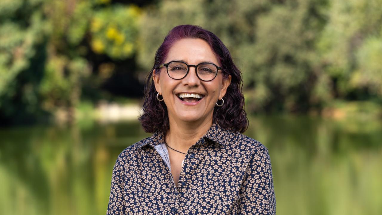 A portrait of Andrea Alfaro standing in front of greenery and joyfully laughing into the camera.