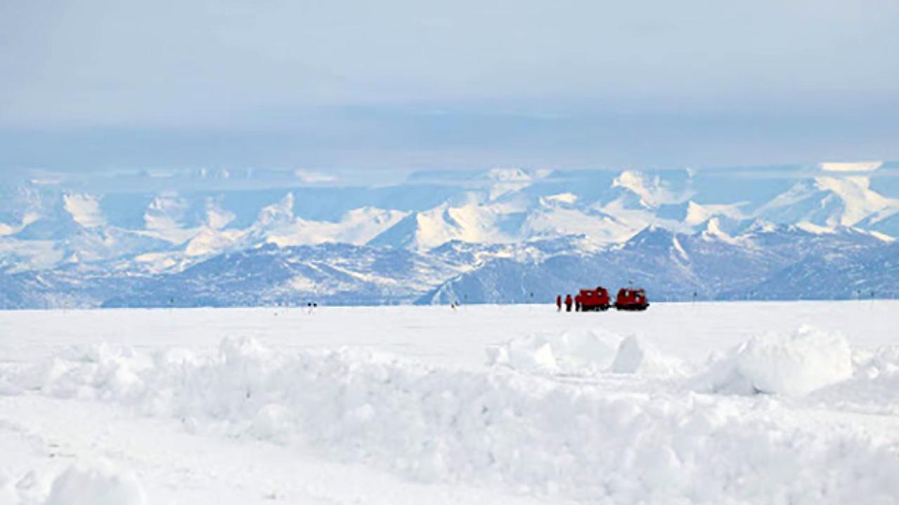 Snowy ground in the foreground and snow covered mountains in the background, against which two bulky red vehicles are visible far off on the horizon, along with several people standing next to the vehicles.