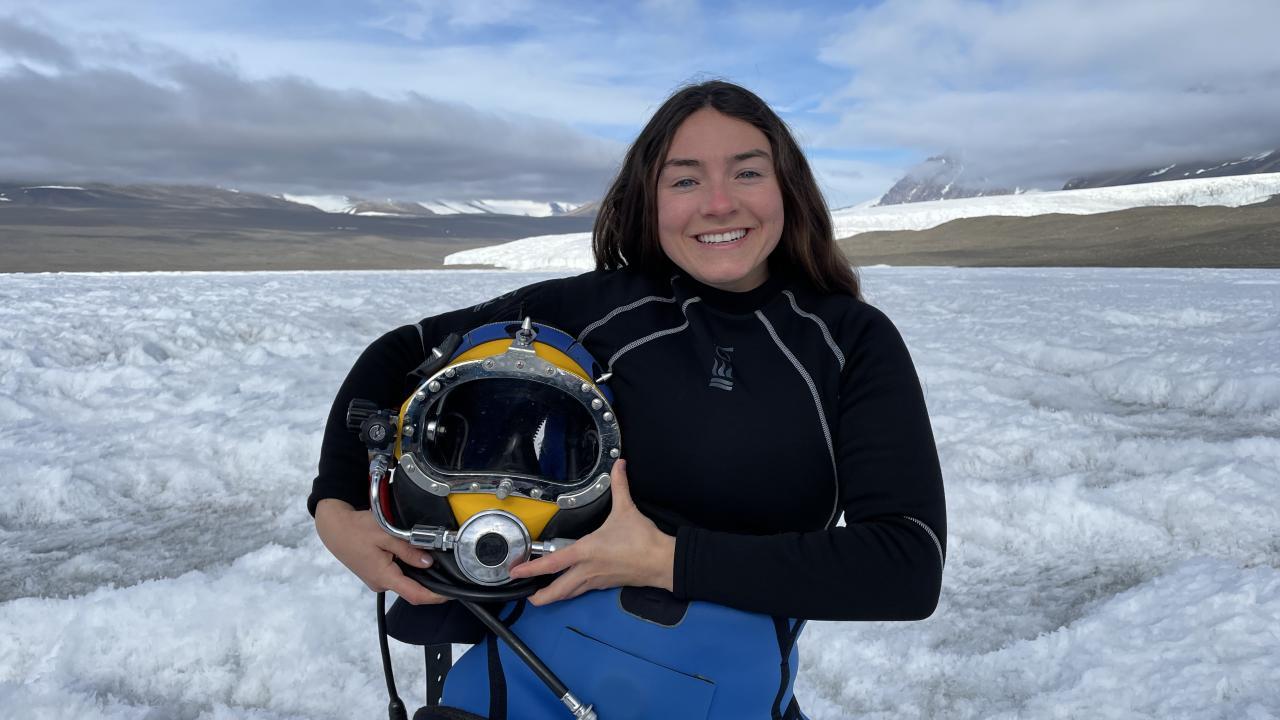 A portrait of Abbey Dias, holding diving gear and standing in front of an expanse of snow.