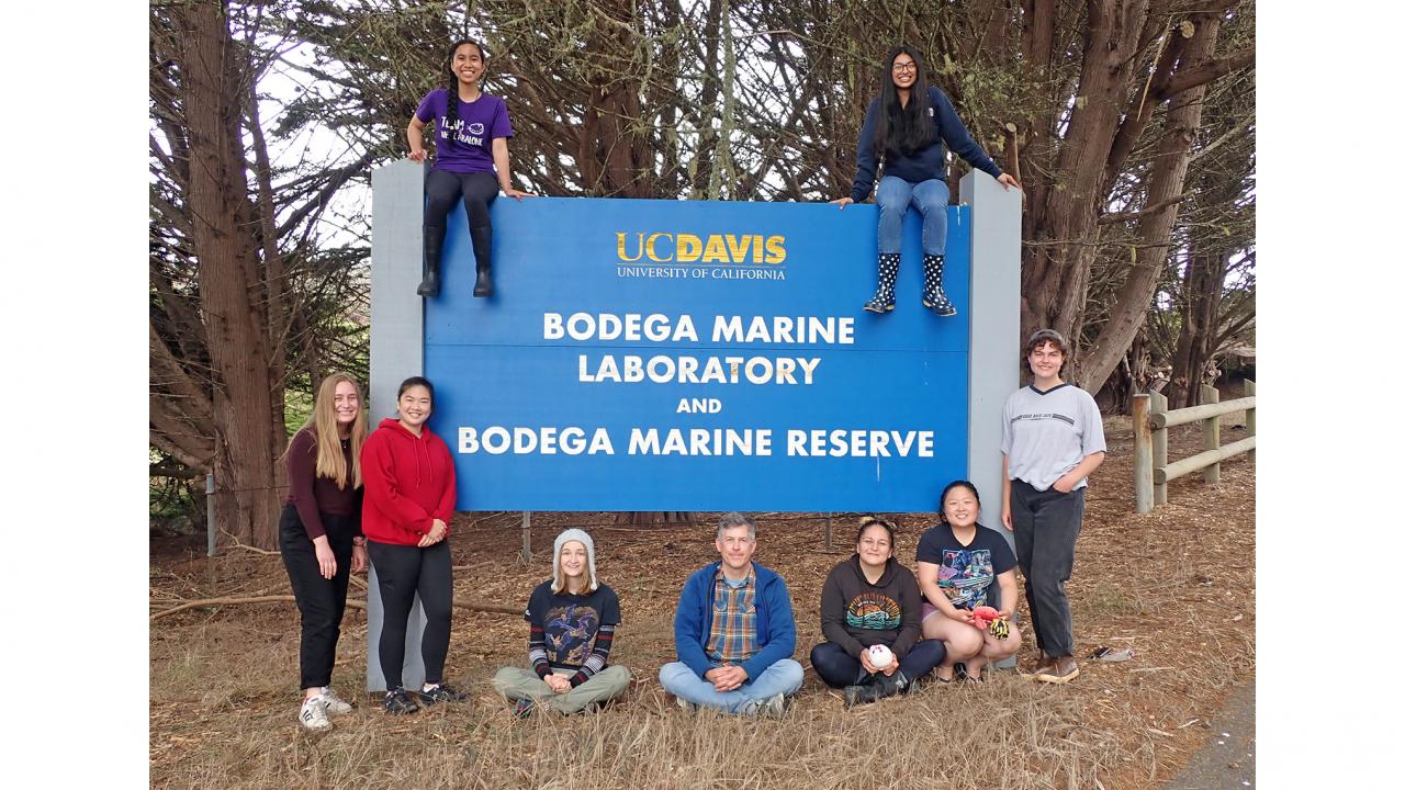 Nine people gathered around a large blue sign that says UC Davis Bodega Marine Laboratory and Bodega Marine Reserve