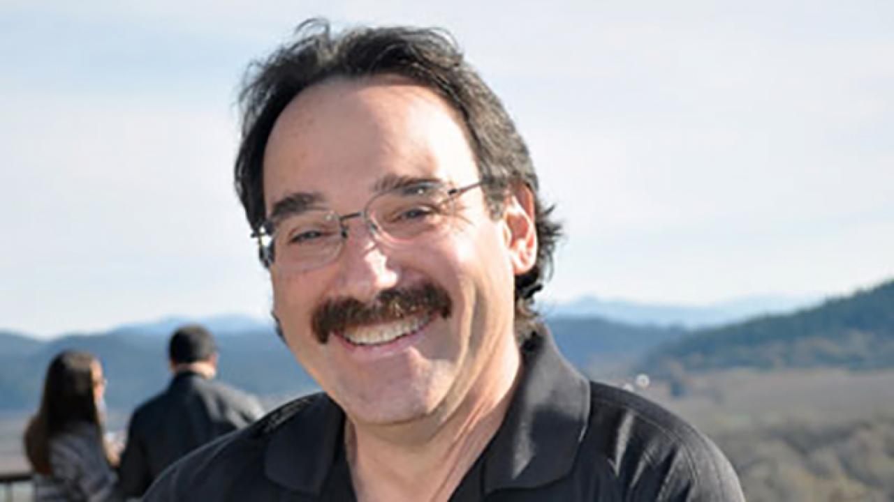 A portrait of a person with dark hair, a thick moustache and glasses smiling at the camera.