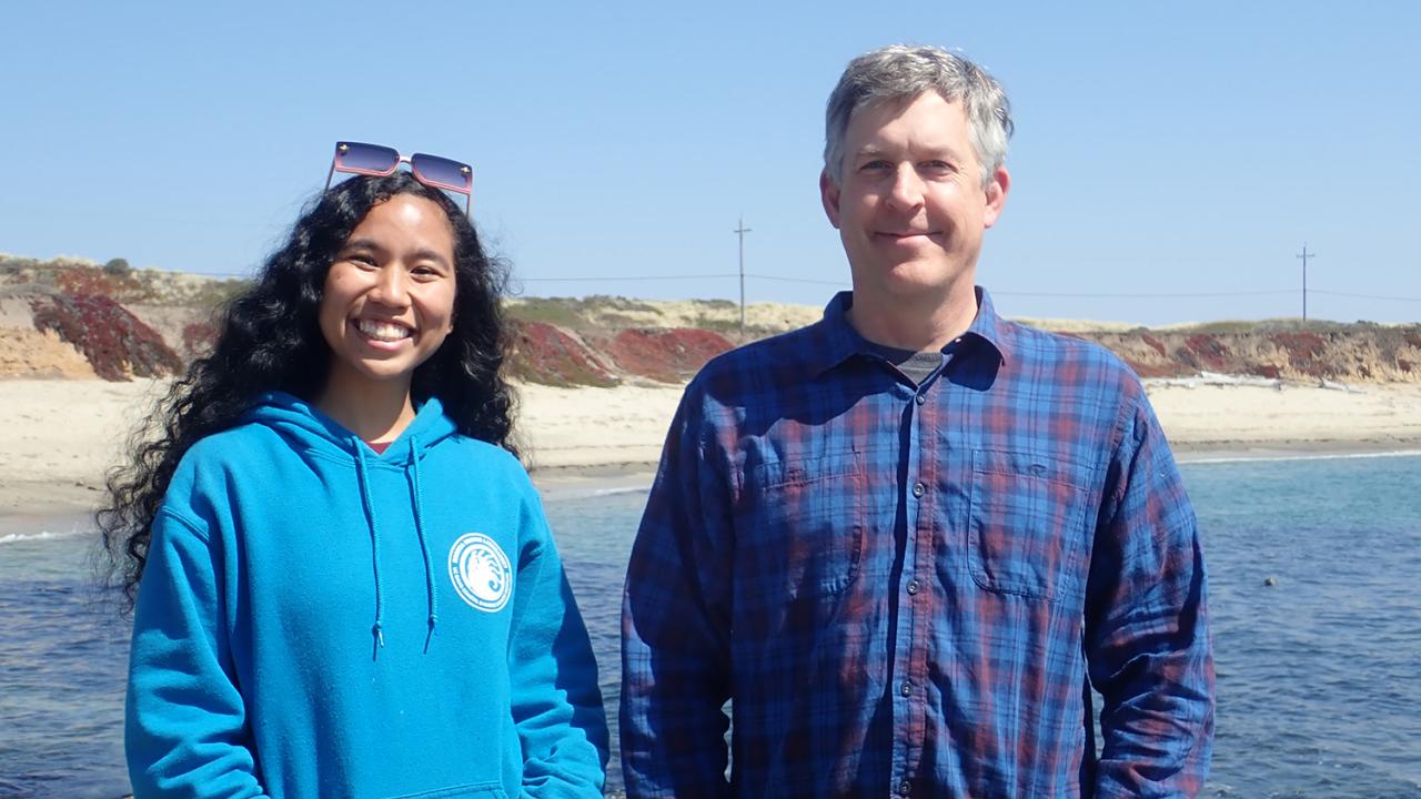 Two people smiling in the sunshine with the beach in the background.