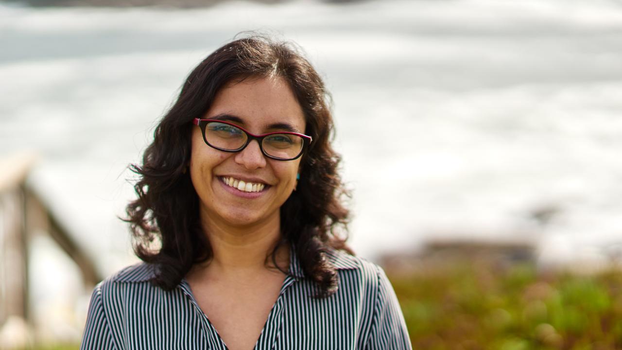 A person with dark curly hair and glasses, smiling at the camera with a coastal scene in the background.