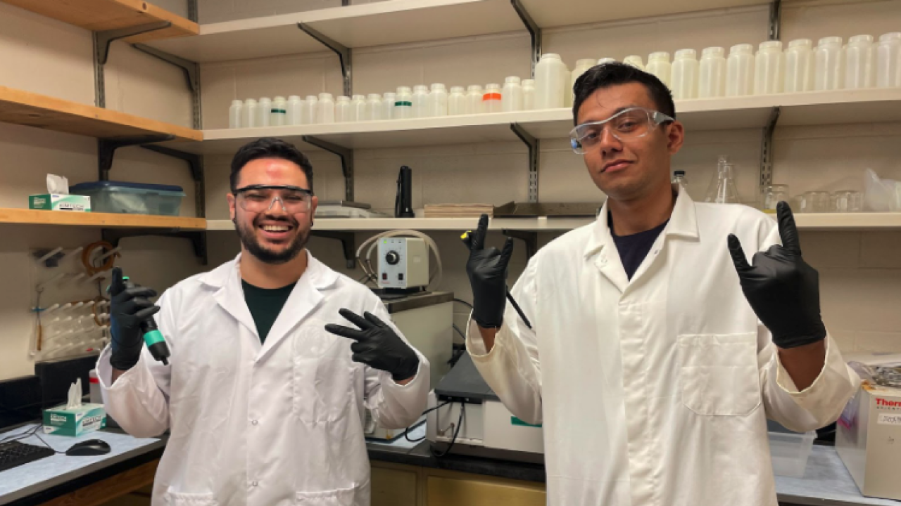 Two people in white coats and safety gear working in a lab