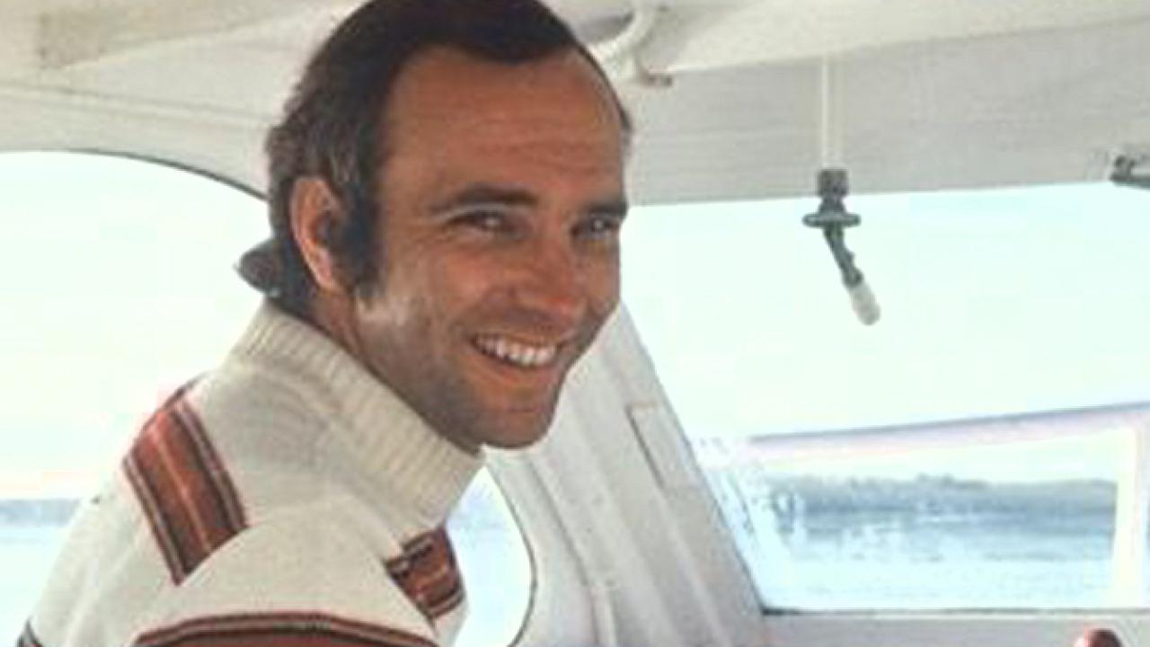 A male with short blond hair, wearing a white and red striped turtleneck sweater and driving a boat.
