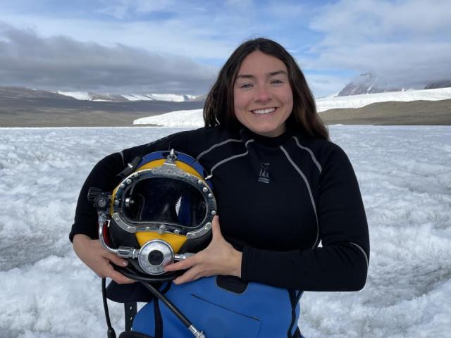 A portrait of Abbey Dias, holding diving gear and standing in front of an expanse of snow.