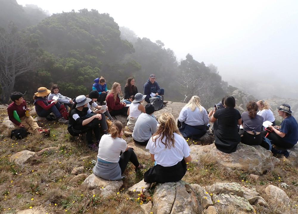 A group of students sitting on rocks on a coastal overlook, deep in discussion.