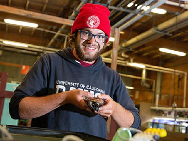 A person in a bright red BML beanie holding up an abalone and smiling at the camera.