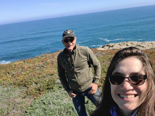 Two people on the coastline with the ocean behind them. In the foreground is a person with long dark hair and black sunglasses, smiling. In the background is a person in a jacket and baseball cap with sunglasses.