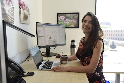 A person in a dark short sleeved shirt with a floral print sits at a desk in front of a laptop and accessory screen, smiling at the camera.