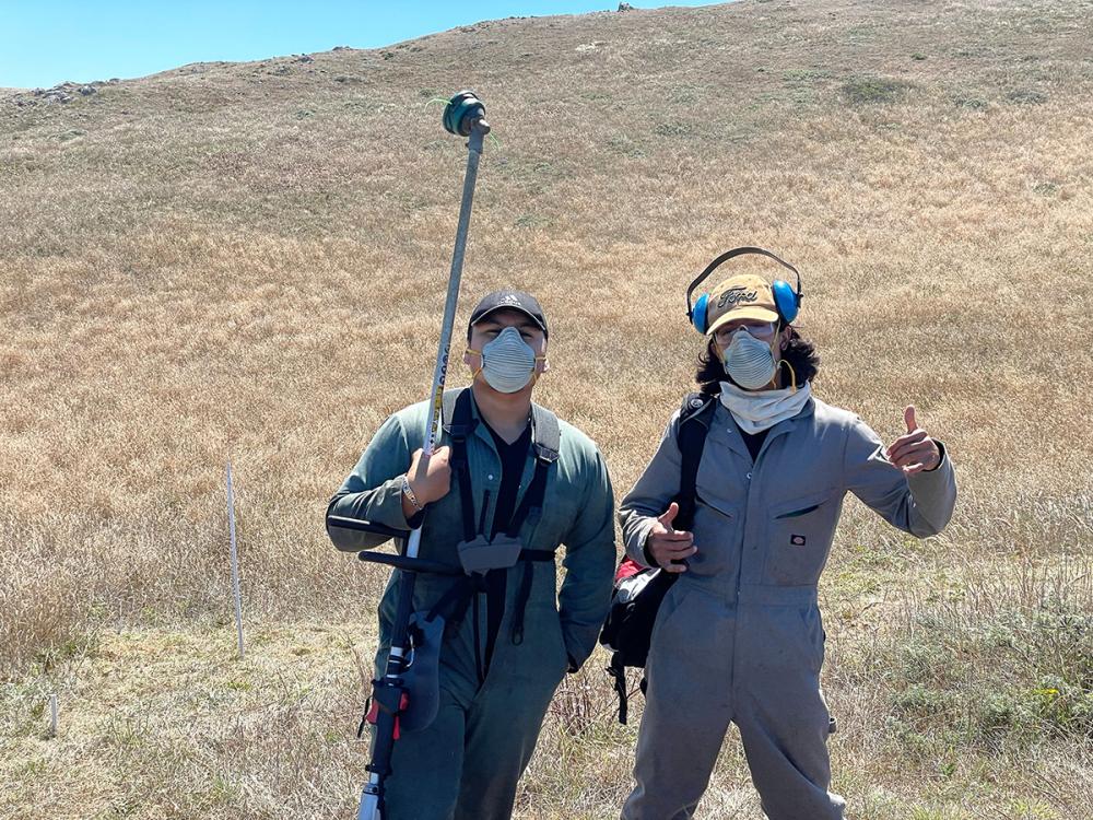 Two people in protective outdoor gear holding handheld mowers in front of a background of coastal prairie