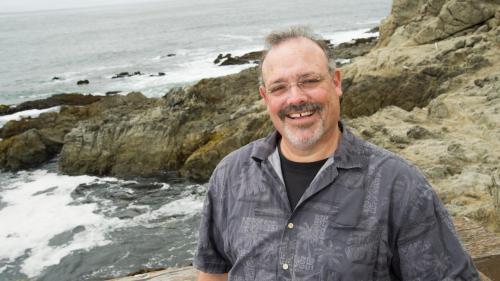 a person in a gray jacket and glasses smiling in front of a rocky coastal background.