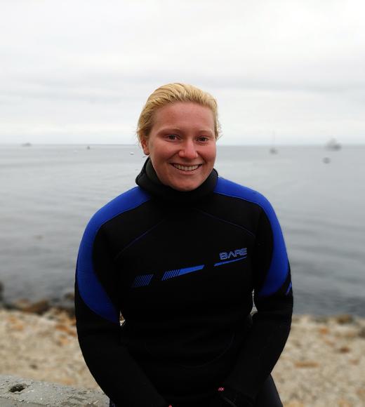 A person in a black and blue wet suite standing in front of a calm ocean coastline.