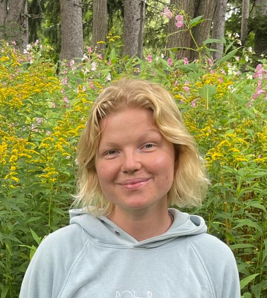A portrait of Grace Callahan standing in front of a wooded area with yellow flowers.