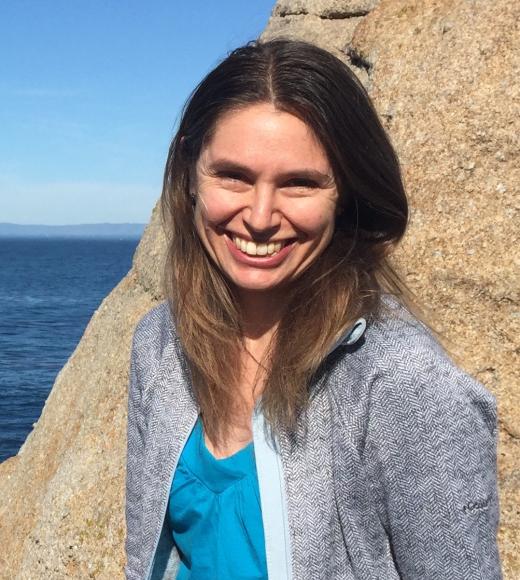 A profile image of a person outside on the coast, with long brown hear and wearing a gray jacket and blue shirt