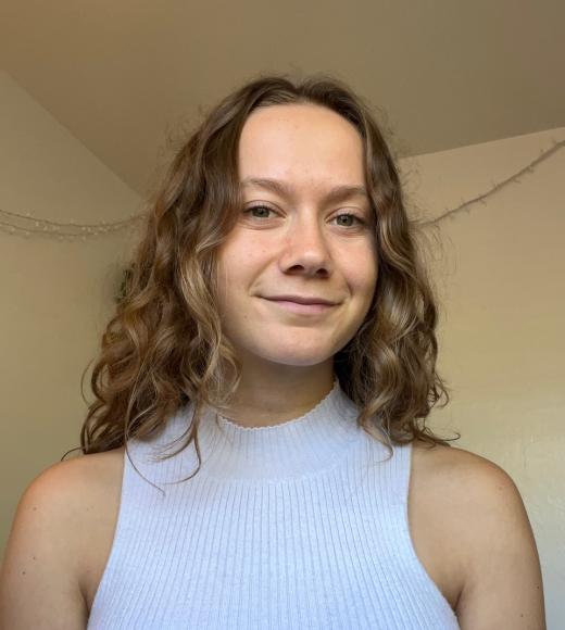 A person with long curly hair wearing a white sleeveless turtleneck and standing in front of a light background