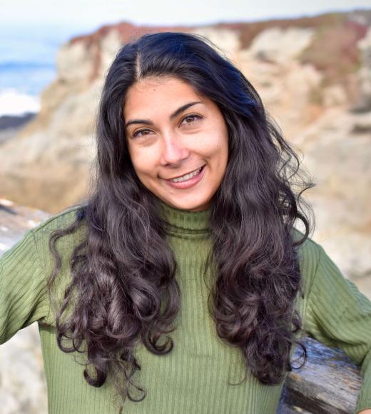 A portrait of Audrey Deutsch standing in front of a coastal background and wearing a high necked green sweater.