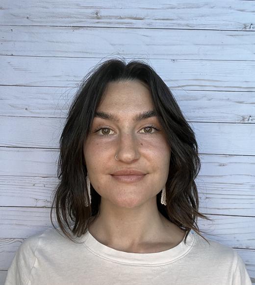 A portrait of a person with dark hair looking straight at the camera with a white wooden slat background