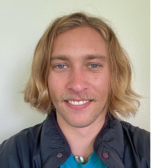 A person with jaw length wavy blonde hair standing in front of a light colored background and looking at the camera