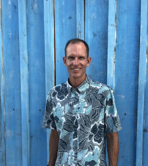 A person with short dark hair wearing a blue shaded hawaiian shirt and standing in front of a blue wood background