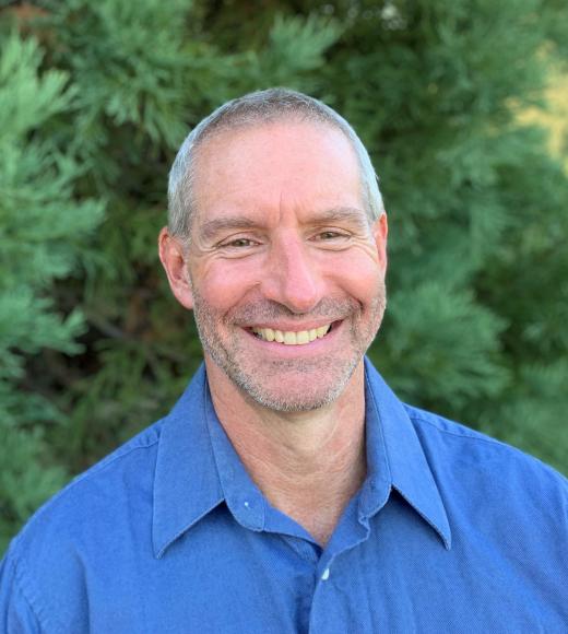 A profile image of a person with short hair wearing a button down blue shirt and standing in front of greenery.