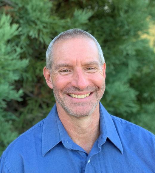 A profile image of a person with short hair wearing a button down blue shirt and standing in front of greenery.