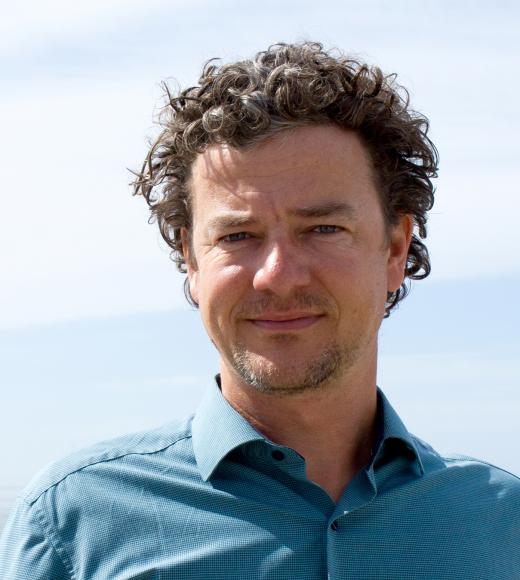 A profile image of a person with short curly hair, wearing a button down shirt and standing in front of a coastline.