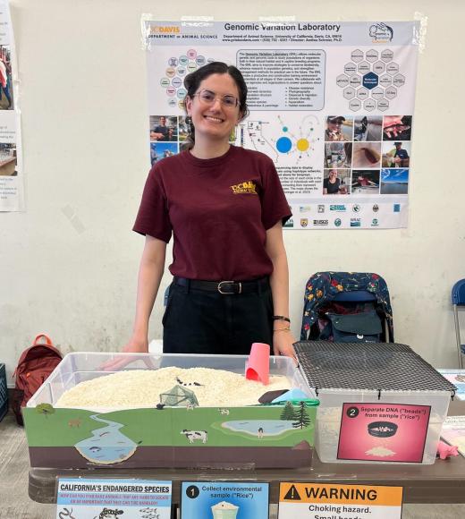 A student standing behind a table that has a large bin filled with rice and strings of beads in it.
