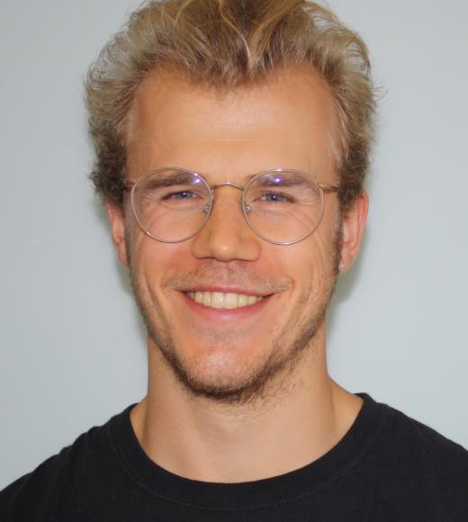 A profile image of a person with short blond hair wearing glasses and a black shirt and standing in front of a white background